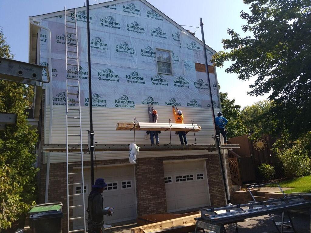 A team of contractors on a pump jack scaffold installs light-colored siding on the second story of a house, working over Kingspan GreenGuard house wrap above a brick garage.