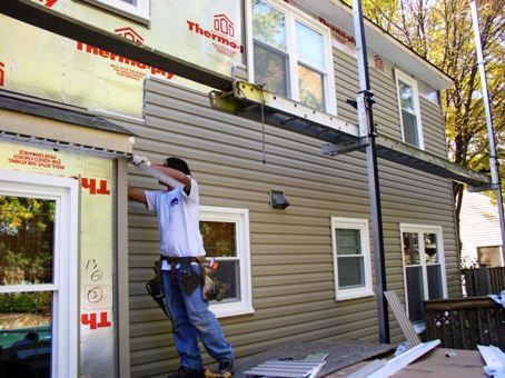 A construction worker in a white t-shirt and tool belt installs tan vinyl siding on the corner of a two-story house. The upper level of the house is partially covered in yellow Thermo-ply protective sheathing, with a metal walkboard and pump jack scaffolding system positioned for higher access. Several white-trimmed windows are visible across the sided and unsided sections of the wall.