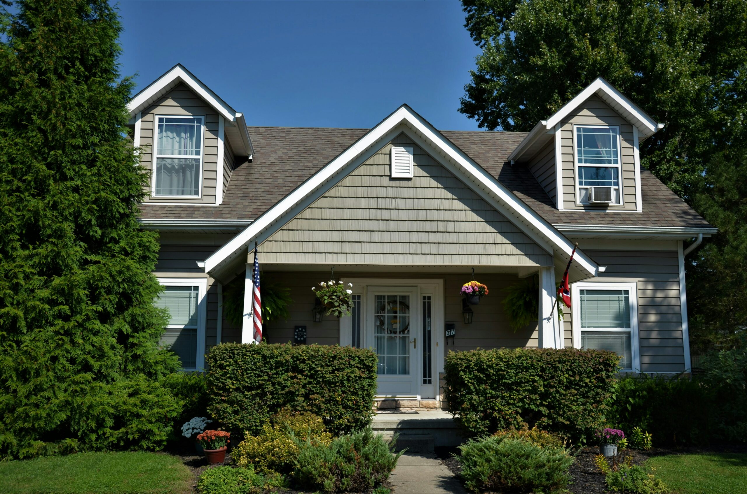 A tan, two-story house with two gabled dormers and a central peaked porch entrance. The home features white trim, a decorative wreath on the front door, and manicured hedges and flower pots lining a concrete walkway under a clear blue sky.