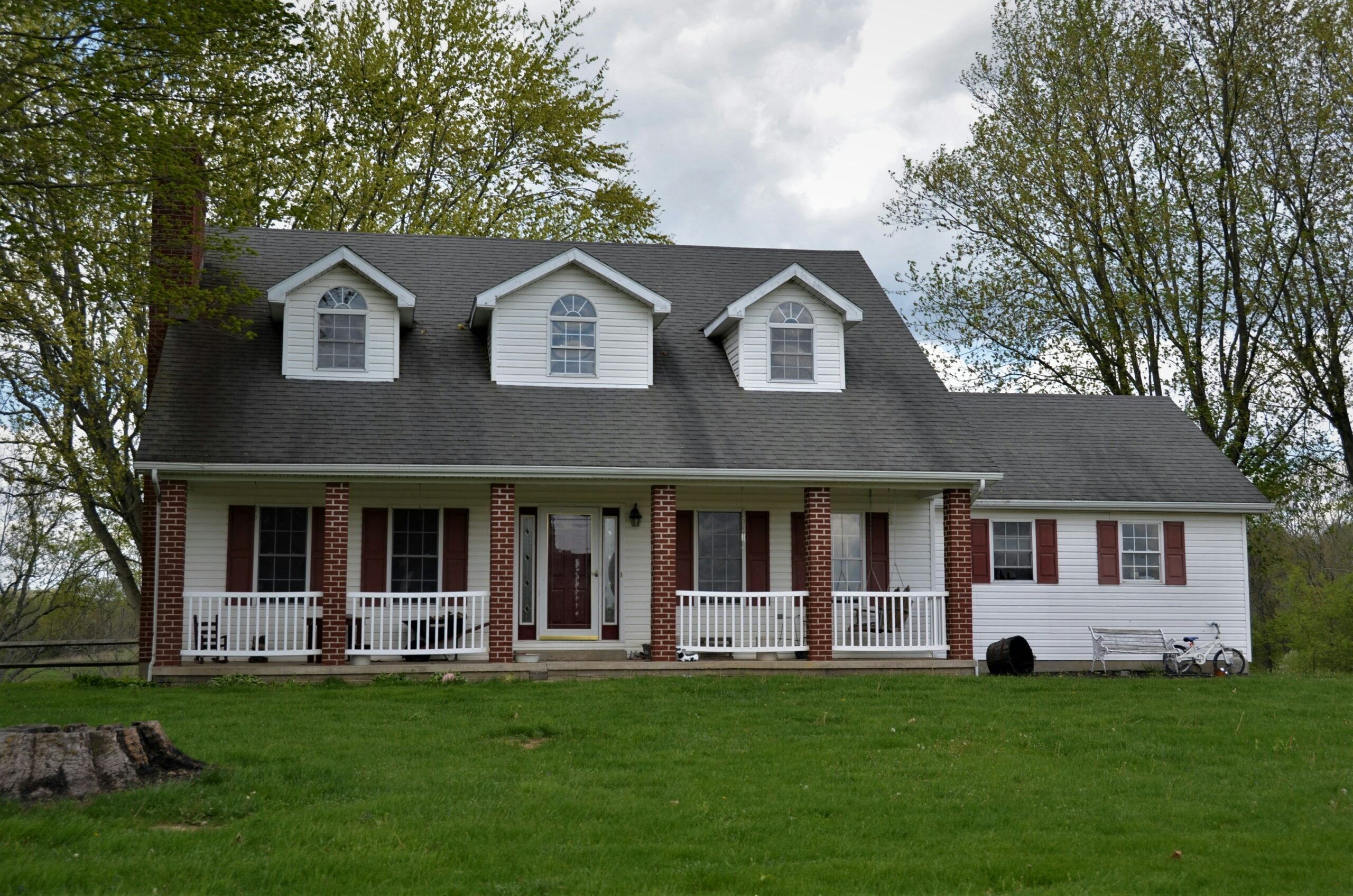 A two-story white farmhouse with three gabled dormers, a dark grey roof, and a front porch supported by red brick pillars. The house features dark red shutters and a matching front door, situated on a green lawn with leafy trees in the background.