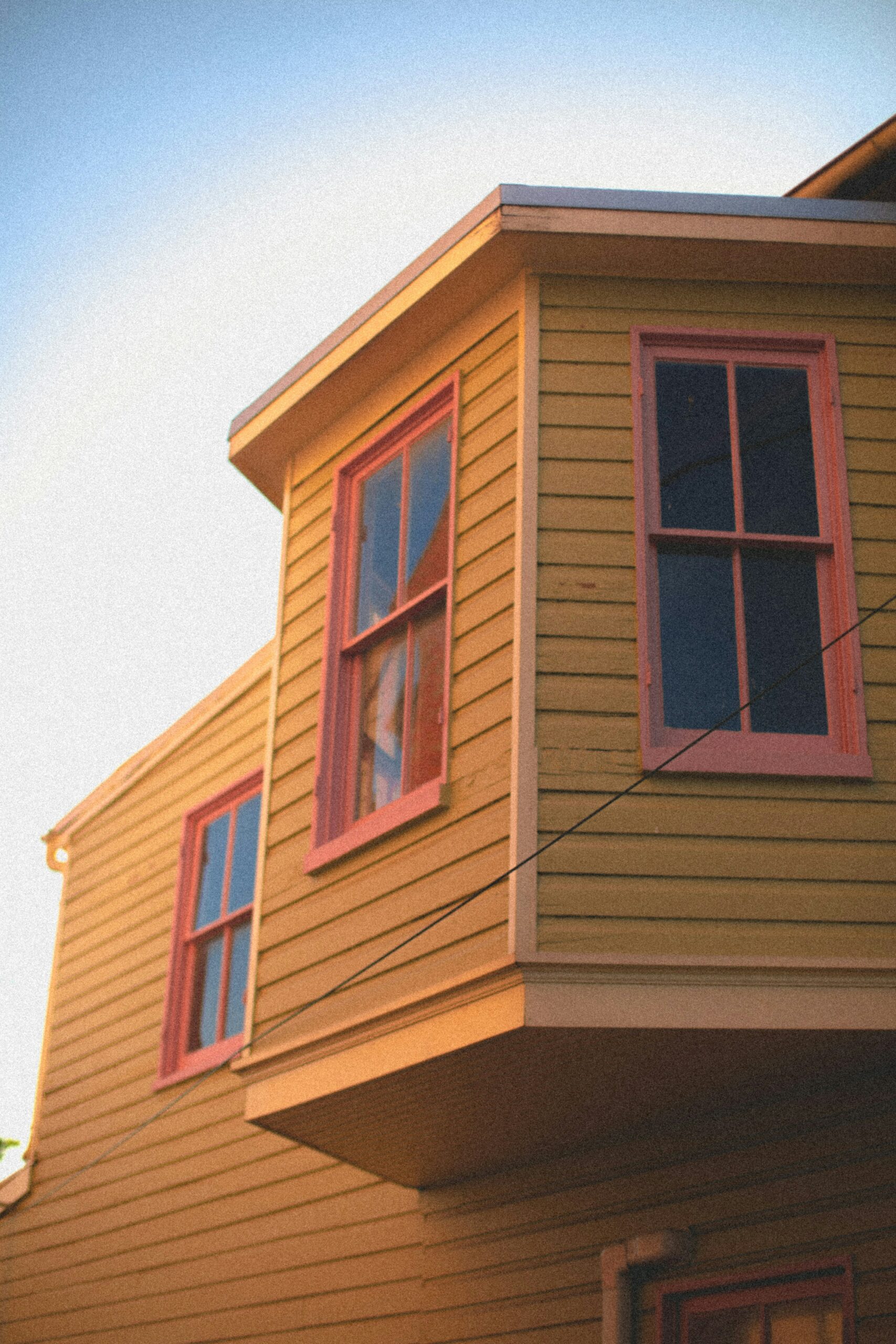 A low-angle shot of a mustard-yellow house featuring horizontal lap siding and a protruding bay window. The windows are framed with contrasting reddish-pink trim, and the warm sunlight casts soft shadows across the textured exterior under a clear sky.